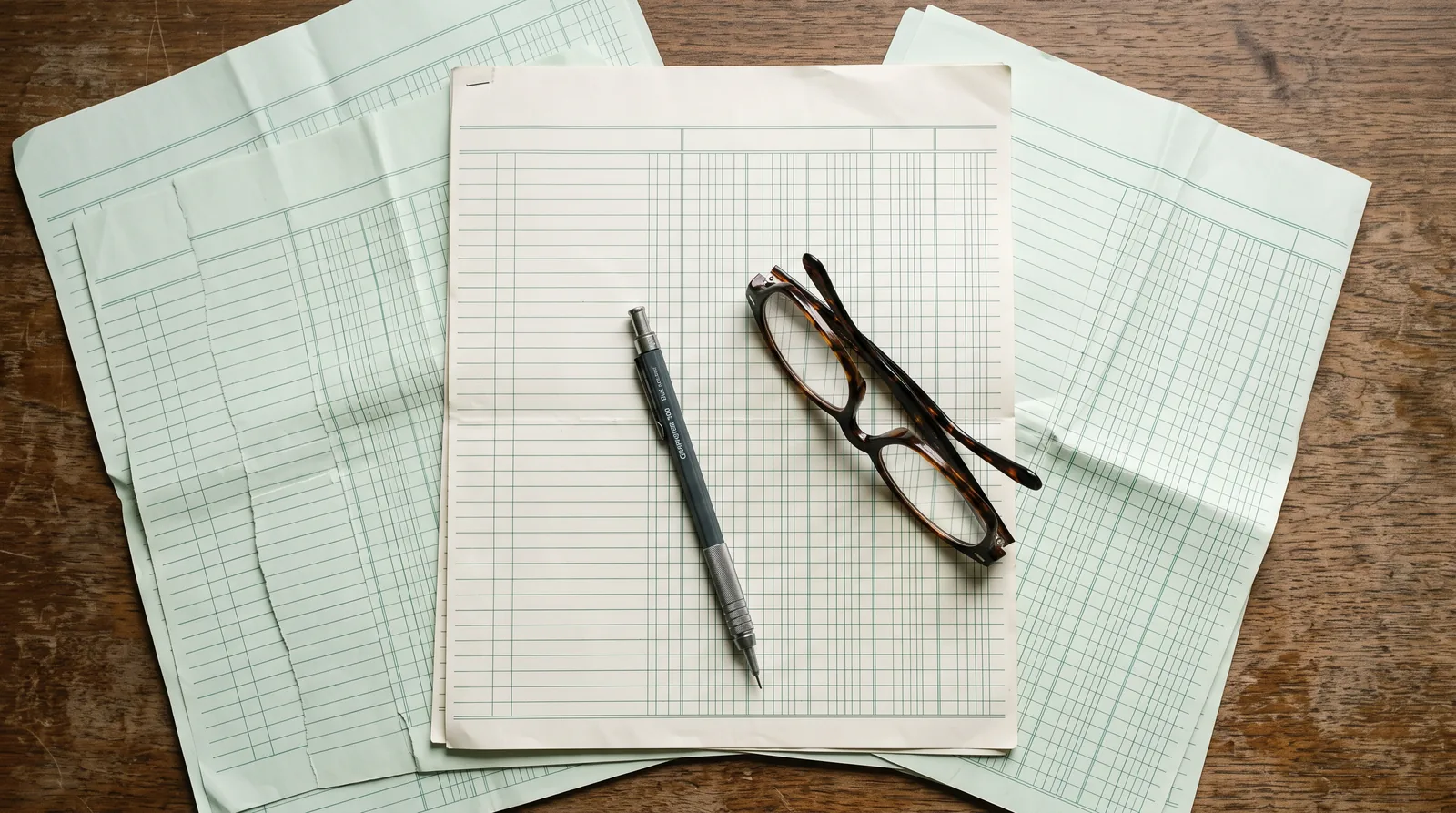 Ruled columnar accounting paper spread across a desk, photographed from overhead with a pencil and reading glasses.