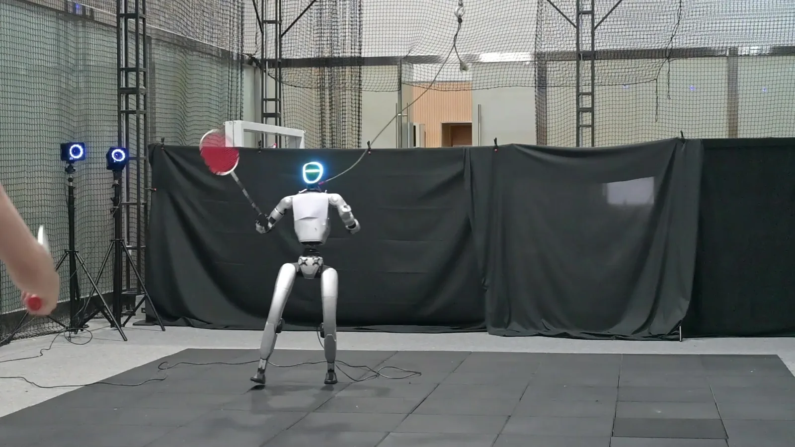 A white humanoid robot with a glowing blue face raises a red badminton racket on a dark practice mat, while a player's arm and racket reach into the frame from the left.