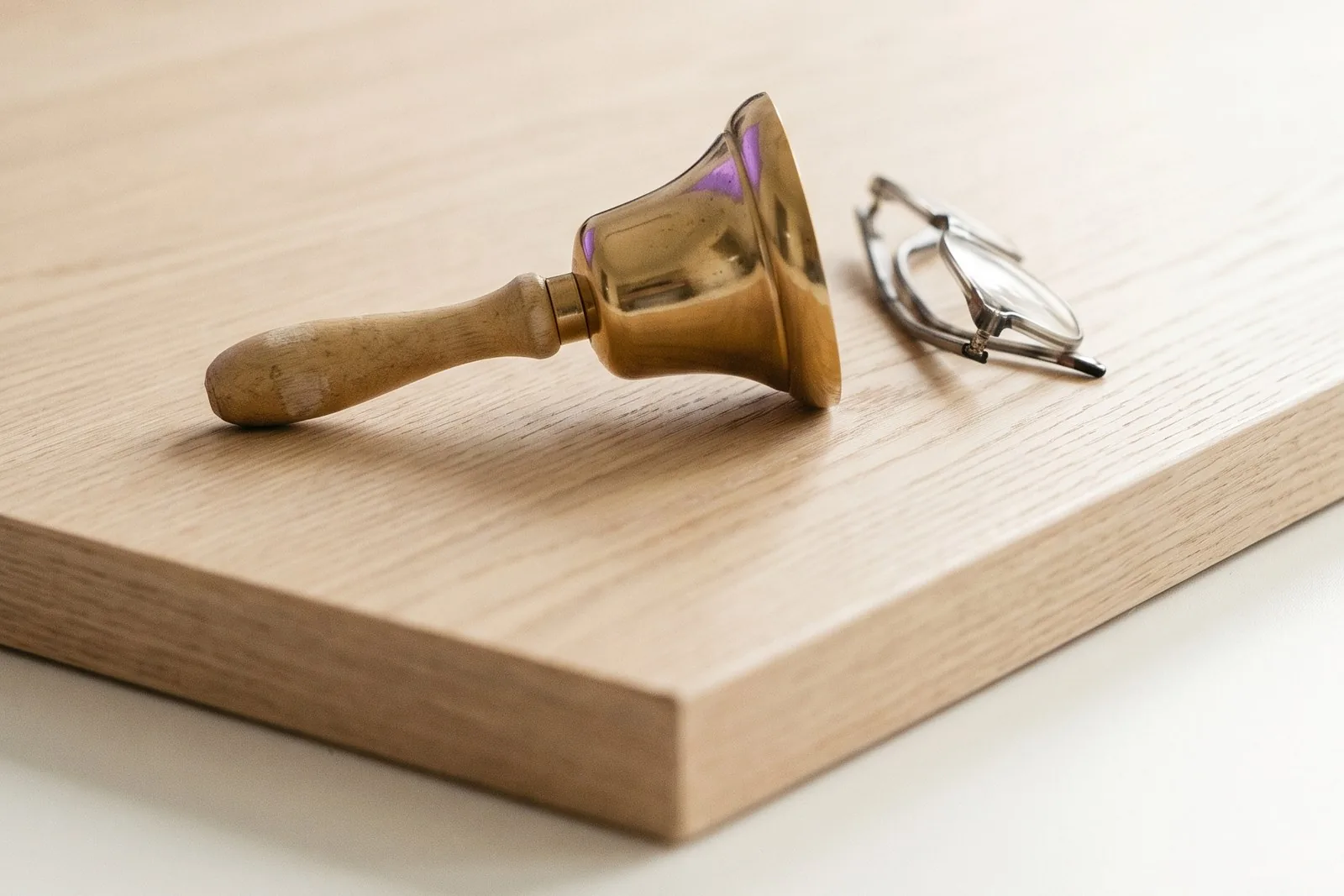 A small brass hand bell lying on its side on a pale oak desk in daylight, beside a folded pair of reading glasses, with the lower half of the frame empty.